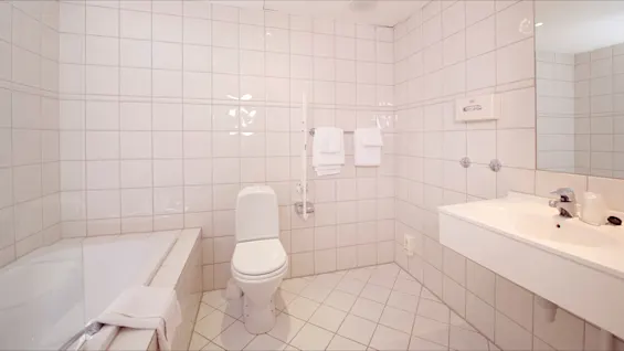 Et lyst flislagt baderom med toalett, badekar, servant og håndklær i et rent, ubeboelig rom.