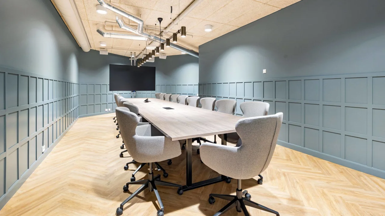 A modern conference room with a long table, chairs, exposed ceiling, and a large screen on the wall.