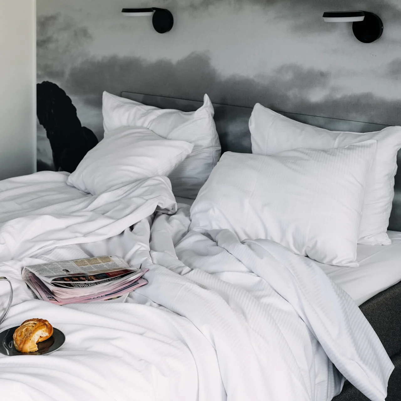 Bed with rumpled white sheets, holding a silver teapot, a cup, a croissant on a plate, and a newspaper. Two pillows rest against a headboard with cloudy wall art.