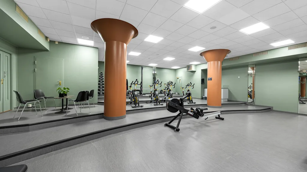 Et romslig hotellgym med treningsapparater, grønne vegger og store oransje søyler.