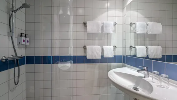 Bruser sidder ved flisevæg med to sæbedispensere og overhængende brusehoved, omgivet af hvide og blå fliser; tilstødende vask har glas under spejlvæg med håndklæder hængende i nærheden.