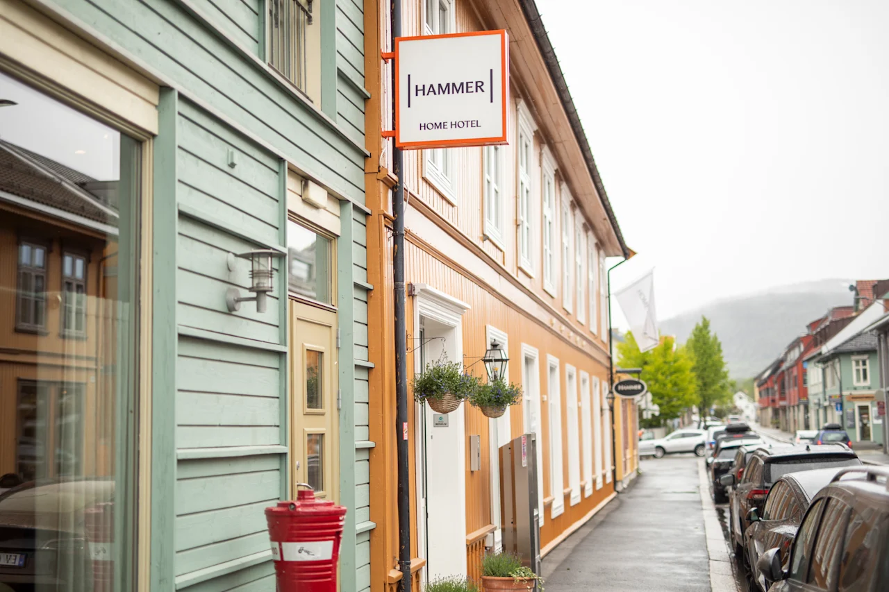 Bygning skilt med teksten "HAMMER HOME HOTEL" på en oransje bygning. Toetasjes bygninger langs en våt gate med parkerte biler. Trær synlige i bakgrunnen.
