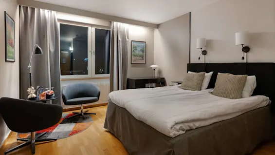 Et hotelværelse med en seng med to puder, to lænestole ved et lille bord, og et vindue med gardiner. Plakater på væggene siger "VÄNERSBORG" og "ALLEPURE."