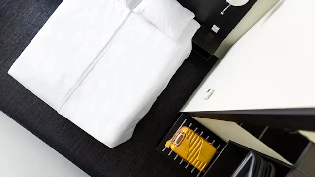 A neatly made white bed sits in a modern room, with a yellow suitcase placed on a rack beside it against a dark carpeted floor.