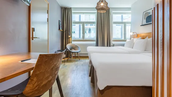A modern hotel room with two beds, a work desk, and a large window, giving a cozy and inviting atmosphere.