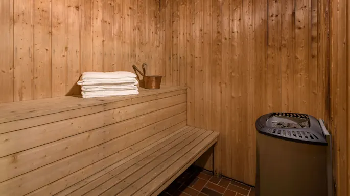 A wooden sauna bench with stacked white towels and a wooden bucket sits next to a metal stove on a tiled floor, surrounded by wooden-paneled walls.