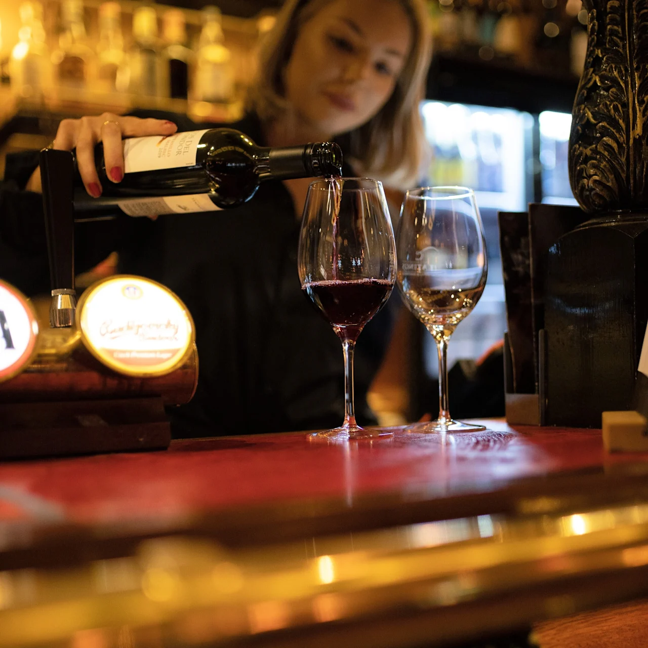 En person häller rödvin från en flaska i ett glas vid en bar. Miljön är varmt upplyst med olika flaskor i bakgrunden.