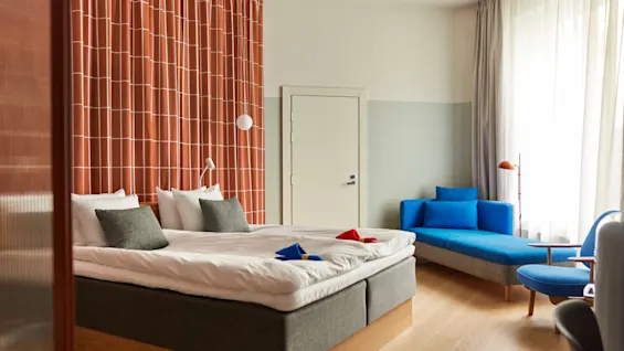 Bright modern hotel room with a double bed, orange patterned curtains, and a blue chaise lounge.