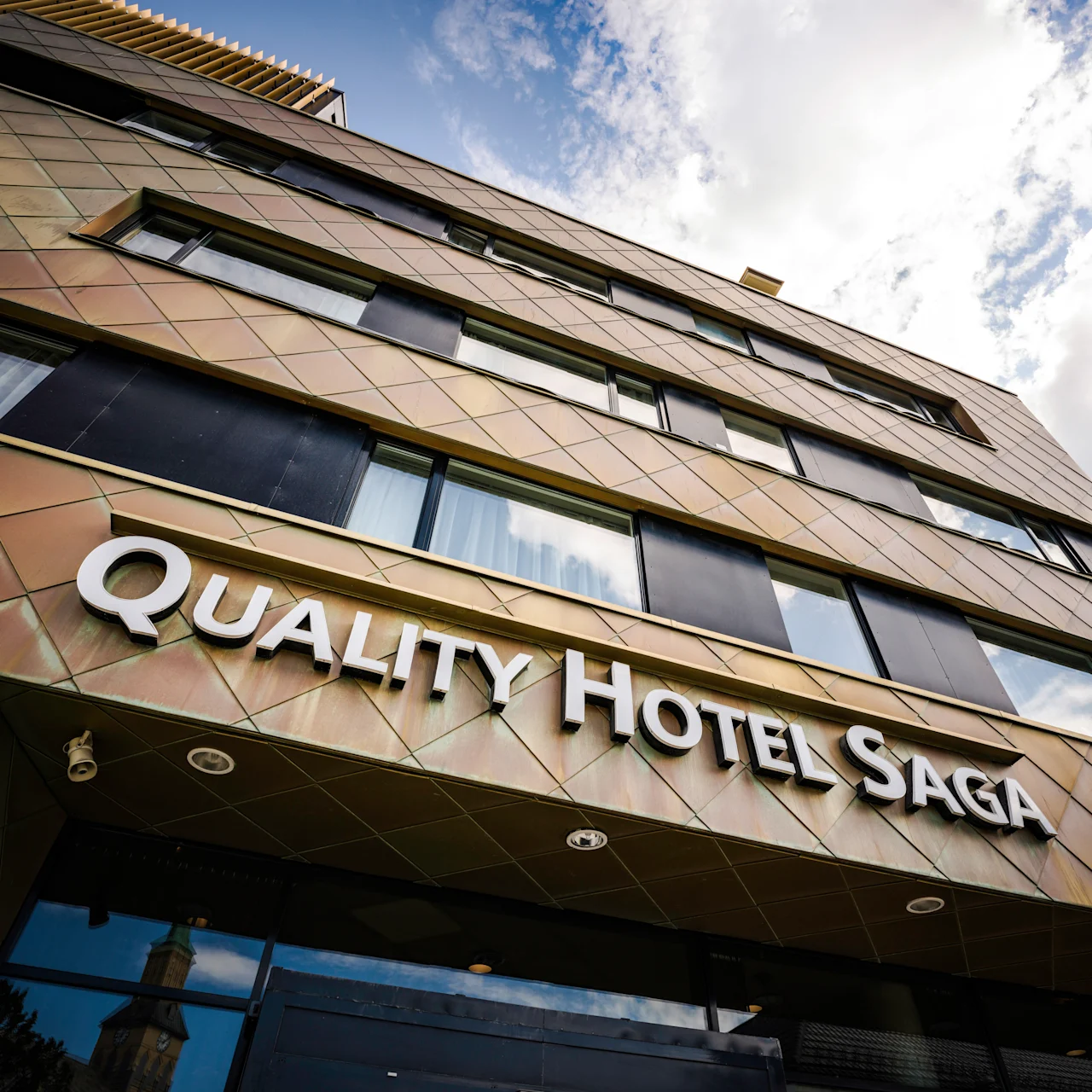 Building features a modern facade with windows; large white letters spell "QUALITY HOTEL SAGA." Surrounding context includes a cloudy sky.