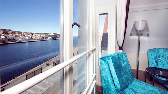 Interior view from a room with turquoise chairs towards an open window showing a waterfront and buildings.