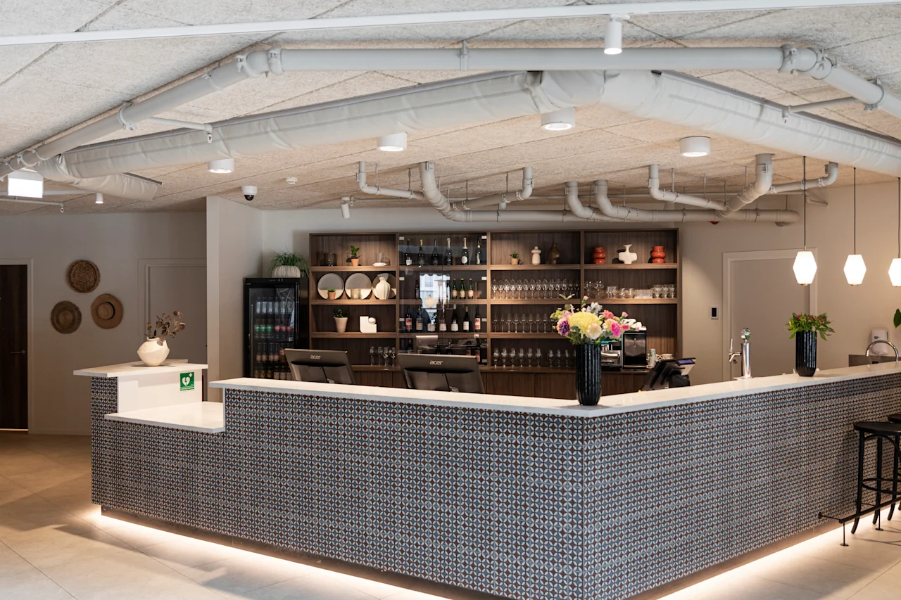 A welcoming reception or bar area with a patterned counter, soft underlighting, and shelves displaying bottles and decorative items.