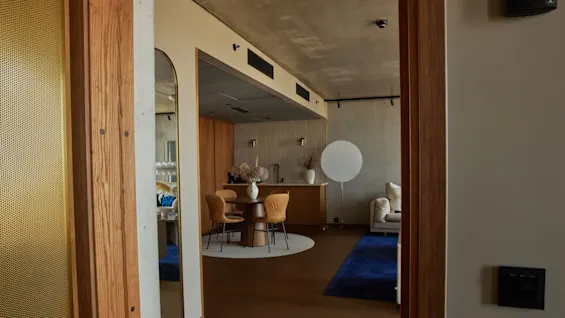 En modern interiör med matbord och stolar, en vas och dekorativa föremål, intill en kokvrå och lounge med soffa och blå matta.