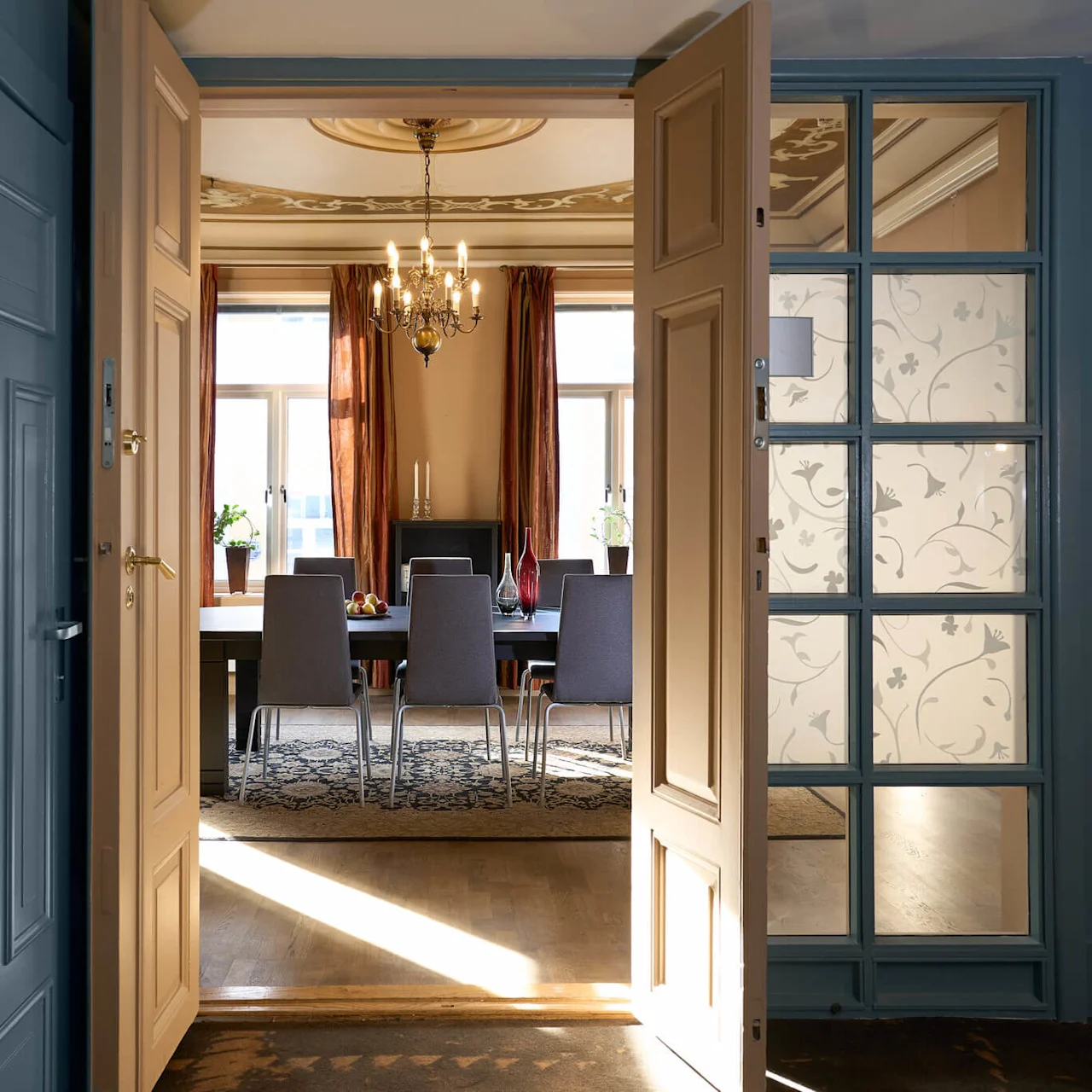 Åbne dobbeltdøre fører til en elegant spisestue med et bord dækket til seks, sollys strømmer ind gennem vinduerne.