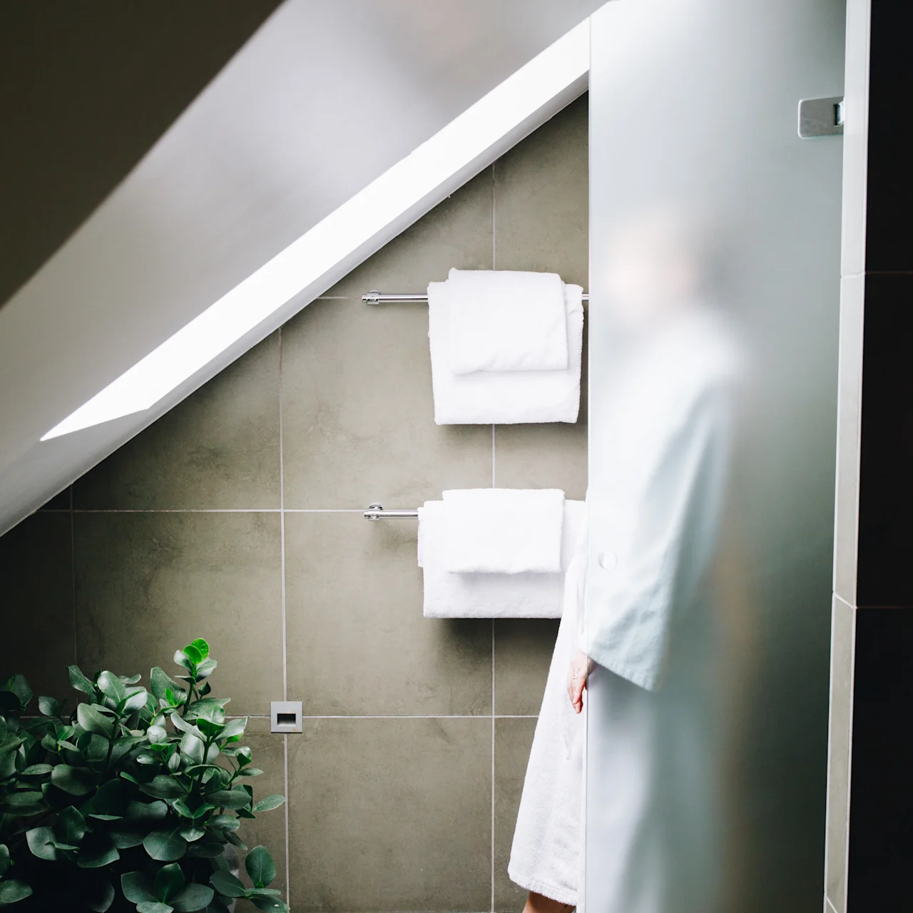 A person stands behind a frosted glass door, wearing a white robe and slippers, in a bathroom with beige tiles, a plant, and two hanging towels.