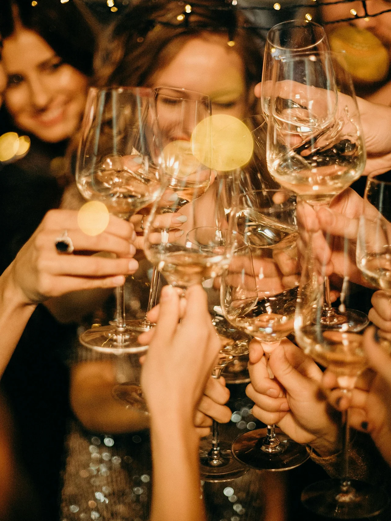 Glasses filled with white wine clink together in celebration, surrounded by smiling people and twinkling string lights in a festive, warm atmosphere.