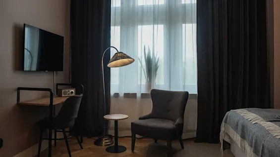 Kodikas sisustus, jossa nojatuoli, pieni pöytä ja lattiavalaisin, lähellä työpöytä, tuoli ja televisio. Suuret tummaverhoiset ikkunat heijastavat pehmeää valoa sängylle.