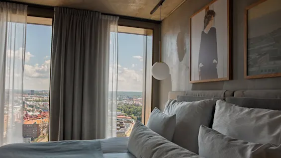 En skickligt bäddad säng med vita kuddar står nära stora fönster. En stadsbild syns genom tunna gardiner. Inramad konst hänger ovanför, och en sfärisk lampa är upphängd i närheten.