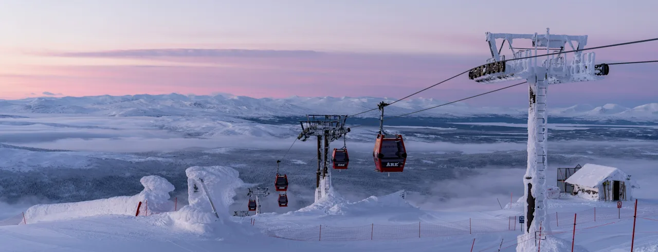 Gondoler stiger opp et snødekt fjell ved soloppgang, med et vidstrakt vinterlandskap og rosa himmel i bakgrunnen. Tekst: ARE.