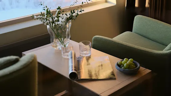 En hyggelig hotelværelsesscene med et sofabord, friske blomster, et magasin, druer og komfortable grønne lænestole ved et vindue.