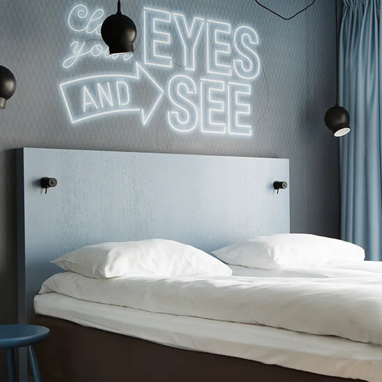 En pent sengt med hvite sengetøy står i et moderne rom med blå detaljer. På veggen står det i neontekst: "Close your eyes and see". To svarte pendellamper henger over.