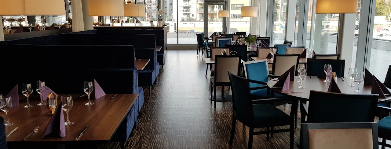 A modern restaurant dining area with set tables, comfortable seating, and large windows offering natural light.