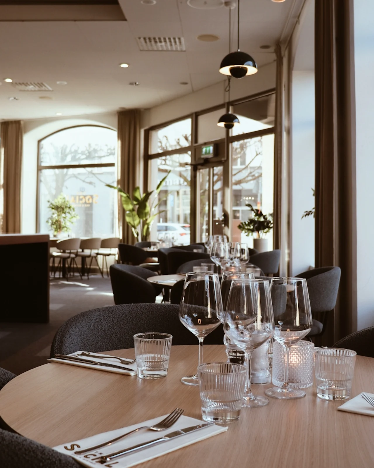 Elegant dining table set with wine glasses and napkins in a modern restaurant; soft lighting creates a warm ambiance. "SICH" text visible on napkin. Large windows reveal trees outside.  