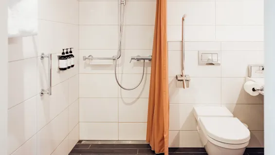 Bruseanlæg med glasflasker, ved siden af et orange forhæng. Ved siden af et moderne toilet med papirholder og metalstøttestang. Hvide flisebelagte vægge, minimalistisk badeværelsesdesign.