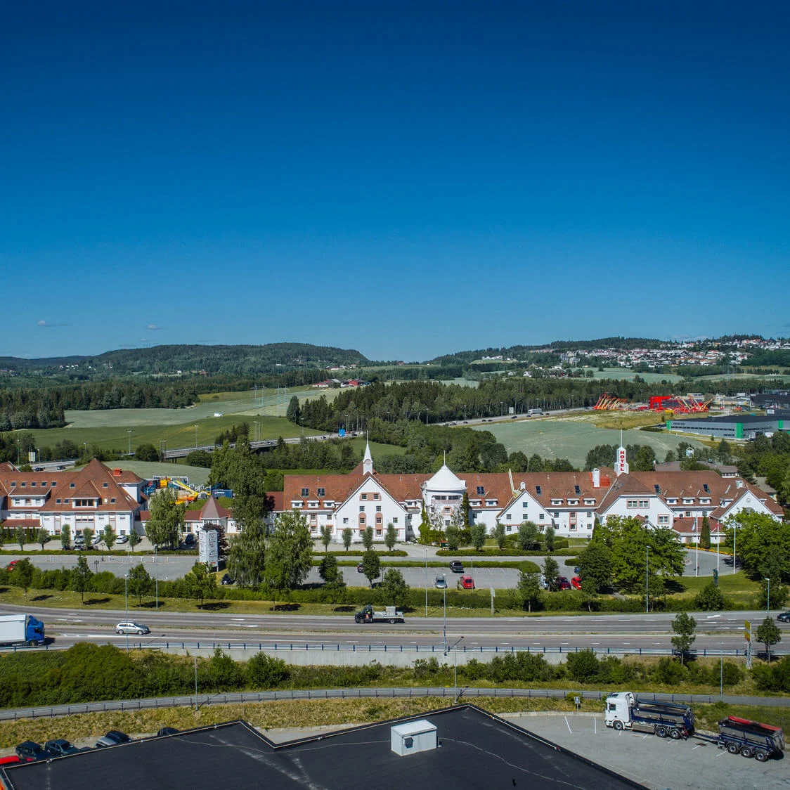 Bygningskompleks med røde tage står blandt frodige grønne marker og træer under en klar blå himmel. Veje med køretøjer omgiver området og giver en naturskøn baggrund.