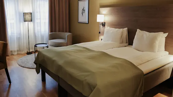 A neatly made double bed with white sheets and green throw stands in a softly lit room. A gray chair, lamp, and table are by the window with closed curtains.