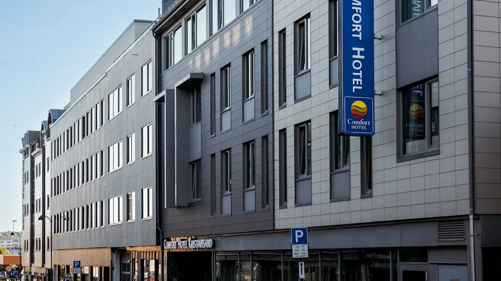 Monikerroksinen hotellirakennus, jossa on sininen kyltti "COMFORT HOTEL" ja "Comfort Hotel Kristiansand." Se sijaitsee kadun varrella, jossa on pysäköityjä autoja. Pysäköintikyltti ilmoittaa "08–17 Man–lør 1t max privat."