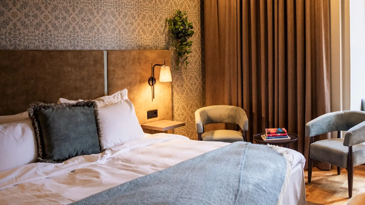 Bed with cushions beside a seating area, under soft lighting. Beige curtains cover a wall, next to a window with a bench adorned with pillows.
