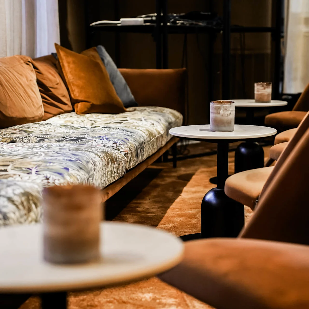 En mönstrad soffa med bruna och grå kuddar ligger intill runda bord med frostade glas i en varmt upplyst, mattbelagd lounge.
