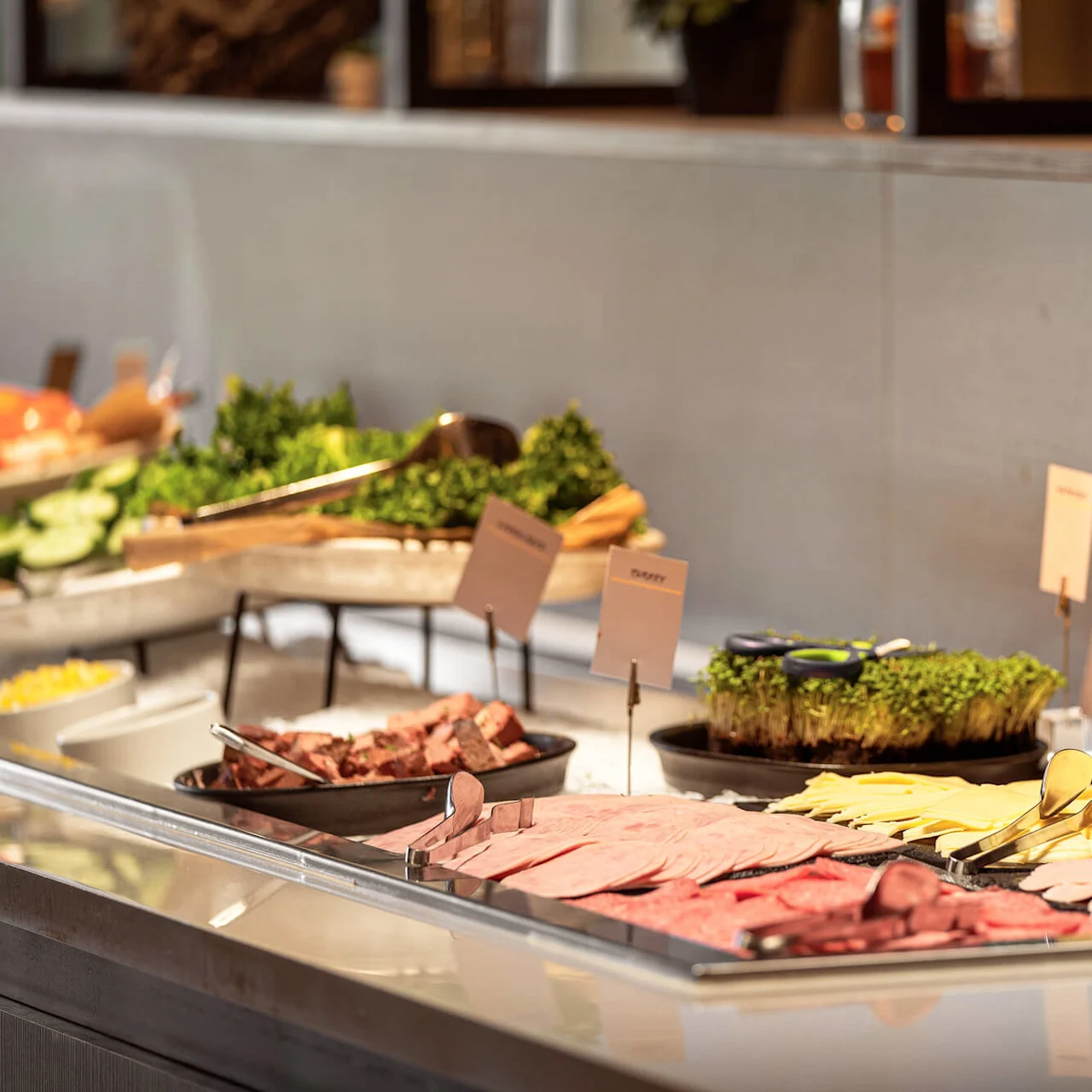 Buffet table displays sliced cold cuts and cheese, accompanied by garnishes. Labels identify the selections. Context is a self-serve dining area with soft lighting.