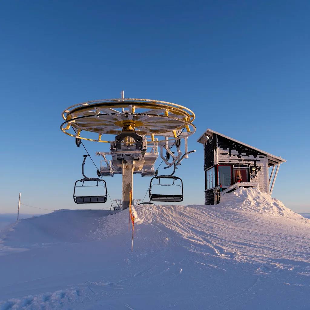 En skidliftstation och en liten stuga på en snötäckt bergstopp under en klarblå himmel, med avlägsna berg och moln.