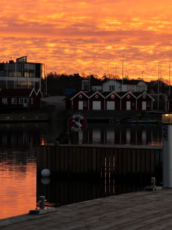 Trebryggen reflekterer en levende oransje solnedgang; små røde bodhus og en moderne glassbygning i bakgrunnen; stille vann omgir, silhuett av master mot fargerik himmel.