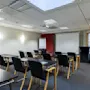Et godt opplyst konferanserom med rader av tomme skrivebord og stoler vendt mot en hvit skjerm, klar for en presentasjon. En flippover står i nærheten, og et høyt bord er plassert på siden.