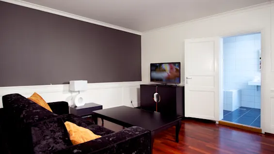 Ett modernt vardagsrum med en svart soffa, TV och soffbord, med utsikt in i ett kaklat badrum.
