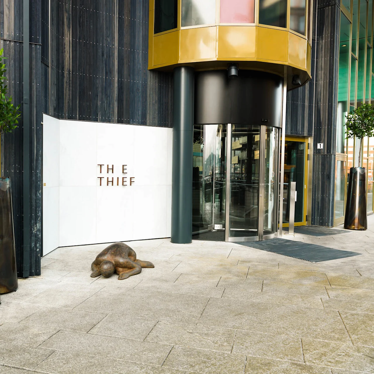 En glassdør som roterer på en moderne bygning med store planter og en skulptur av et liggende dyr. Tekst på veggen lyder "THE THIEF".