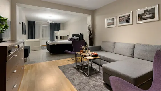 En grå hjørnesofa står i en stue vendt mod et lille bord med snacks, ved siden af et soveværelse med en seng. Indrammede fotografier pryder de beige vægge.