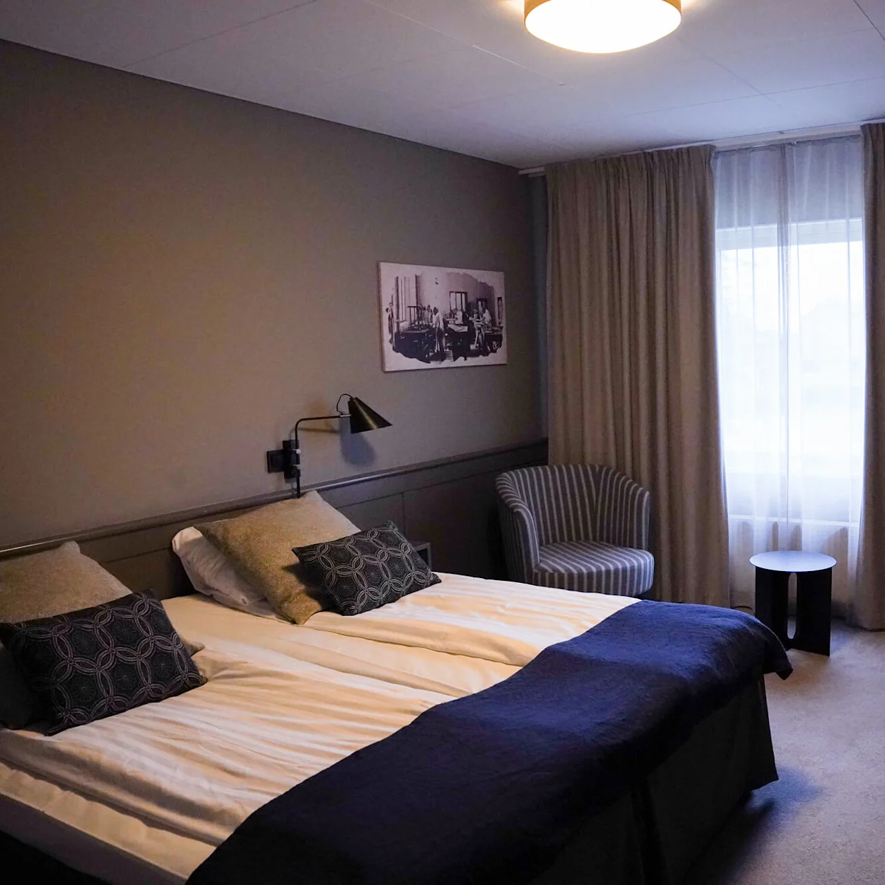 A tidy hotel room with twin beds, softly lit with wall sconces and overhead lighting, featuring a desk, chair, and framed wall art.