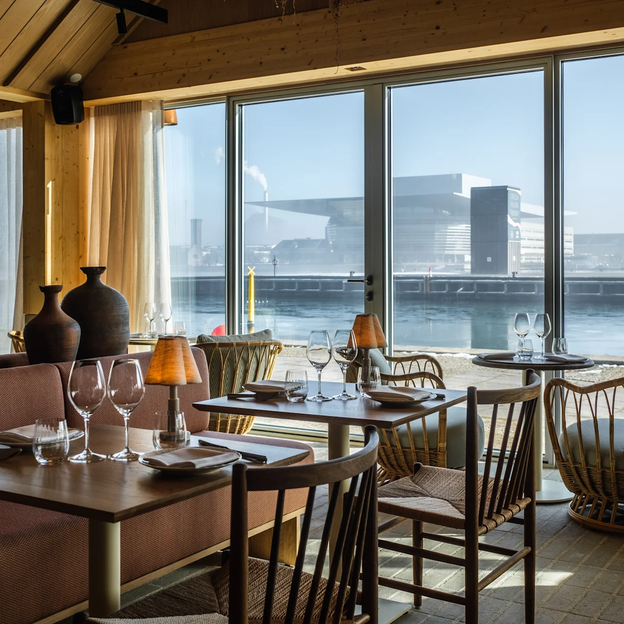Dining tables are set with glassware and napkins, in a cozy restaurant with wooden decor. Large windows reveal a waterfront view with a modern building on a sunny day.