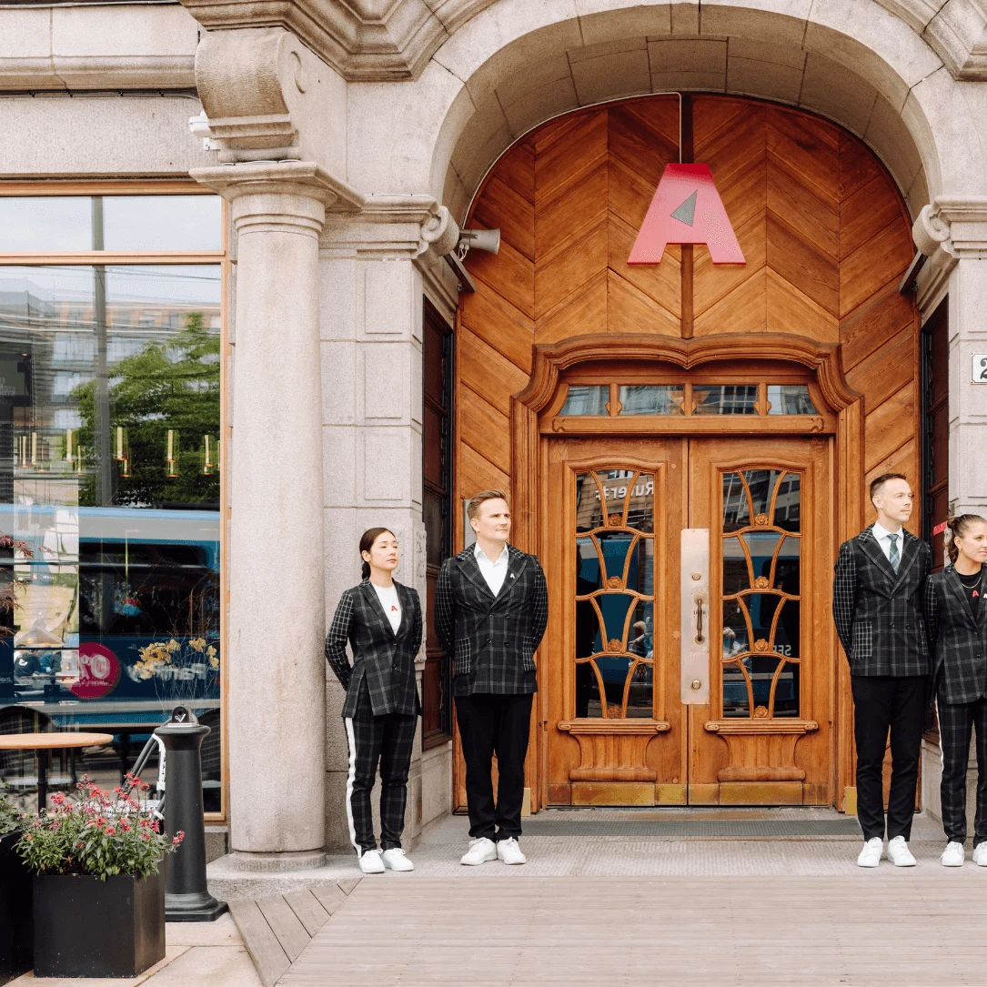 Four people stand outside a large wooden door with an arch, wearing matching plaid suits. There is a large red letter "A" above the door. Nearby, glass windows reflect a busy street. A plaque shows the number "2."