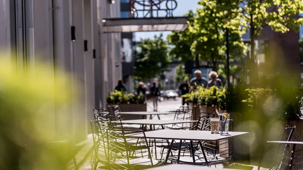 Udendørs metalkaféborde med glasdrikke, omgivet af grønne planter, og et par mennesker, der går langs en solbeskinnet gade under en delvis skyet himmel.