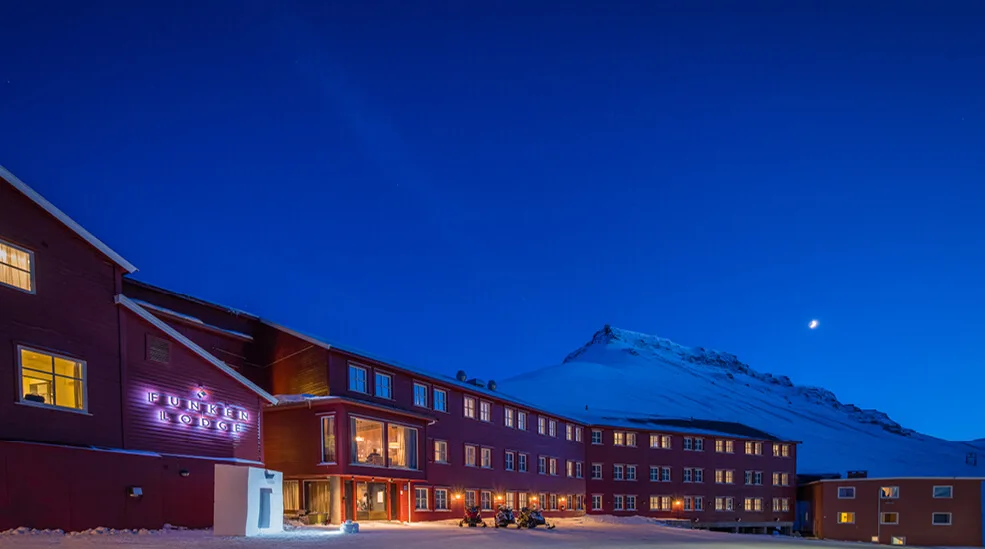 Punainen huvila, jossa on loistavat ikkunat ja neonmerkki "FUNKEN LODGE", sijaitsee hämärän taivaan alla vuoren ja kuunsirpun taustalla.