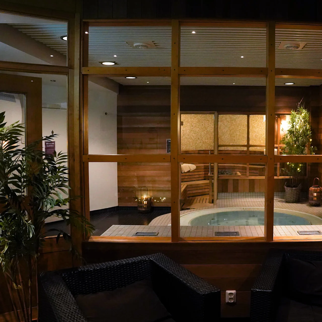 A dimly lit spa area with a Jacuzzi and sauna visible through glass panels, accompanied by indoor plants and soft lighting.