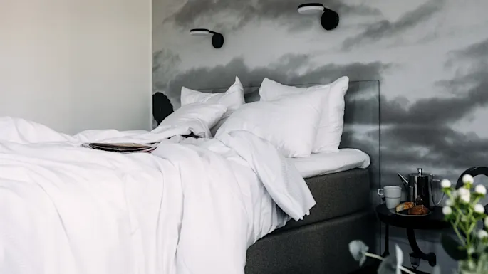 Bed with white linens and pillows is unmade, suggesting recent use. Nearby, a nightstand holds a teapot, cup, croissant, and flowers. Mural of clouds adorns the wall.