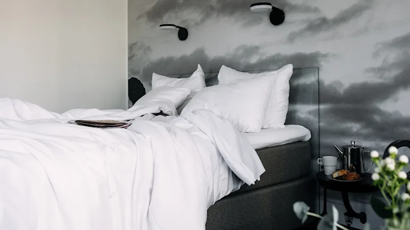 Bed with white linens and pillows is unmade, suggesting recent use. Nearby, a nightstand holds a teapot, cup, croissant, and flowers. Mural of clouds adorns the wall.