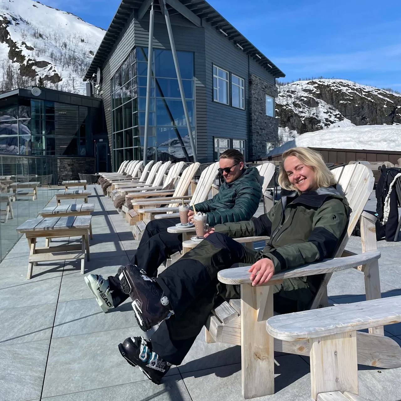 Two people in ski gear relax on wooden chairs, sipping drinks outside a modern building. Snow-covered mountains surround them under a clear blue sky.