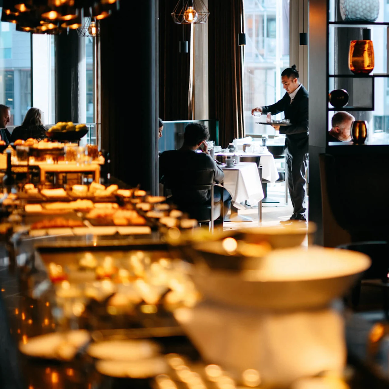 En servitør serverer kunder ved bord i en varmt opplyst restaurant, med dekorative hyller og ulike retter på et buffébord i forgrunnen.
