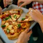 En pizza med smält ost och basilika delas av flera händer från en låda, omgiven av människor i en avslappnad inomhusmiljö.